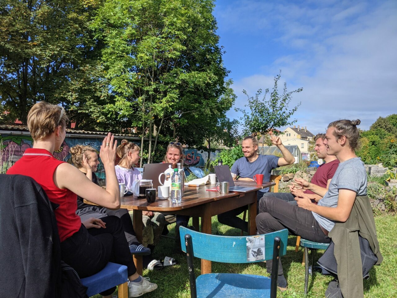 

                          Mehrere junge, weiße Menschen sitzen an einem Tisch unter freiem Himmel, einige heben die Hand zur Abstimmung.

                          