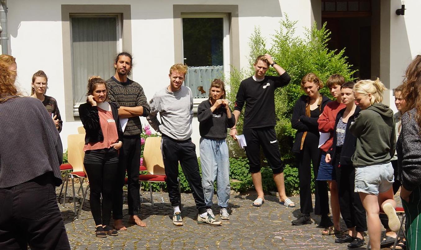 

                          Eine Gruppe junger Leute steht in einem Halbkreis vor einem Haus unter freiem Himmel. 

                          