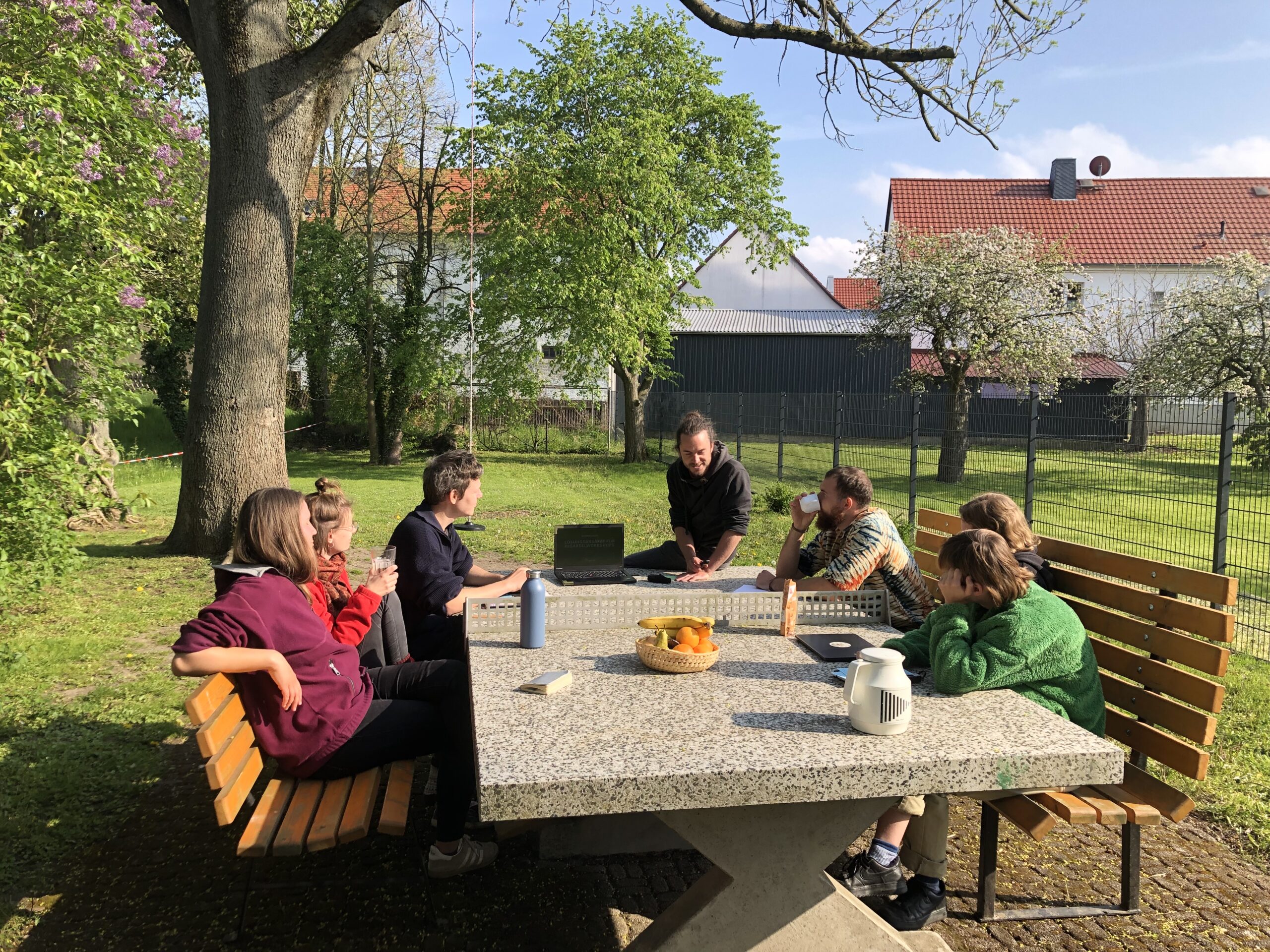 

                          Sieben weiße, junge Menschen sitzen unter freiem Himmel um eine Tischtennisplatte herum und hören einer der Personen zu.

                          